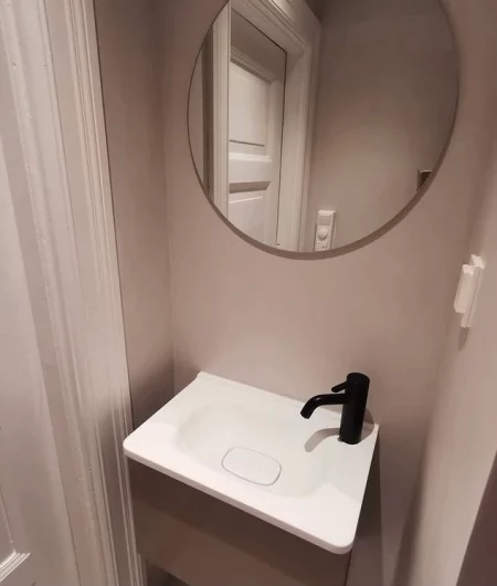 Moderne mikrosement bad med liten servant, svart kran og rundt speil mot lys vegg.